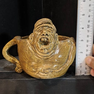 以下が含まれることがあります： 茶色の釉薬がかかった、人の形をしたセラミック製のマグカップ。マグカップには持ち手と広い開口部があります。人の顔は、大きく開いた口と閉じられた目で誇張されています。