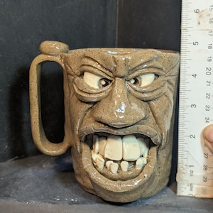 May include: A stoneware mug with a sculpted face design. The mug is tan with a handle and a detailed face with open mouth and teeth. The mug measures approximately 4 inches in height.