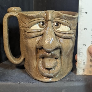May include: A handmade ceramic mug with a sculpted face design. The mug is tan with a handle and features a nose, eyes, and a tongue. The mug is approximately 4 inches tall.