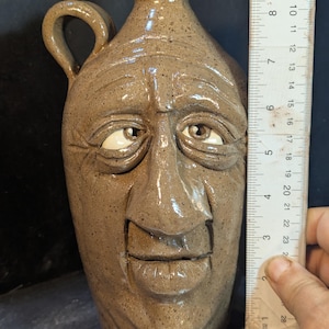 May include: A handmade, brown ceramic jug with a face sculpted on the front. The jug has a handle and a spout. The face has detailed features, including eyes, a nose, and a mouth. The jug is approximately 20 cm tall.