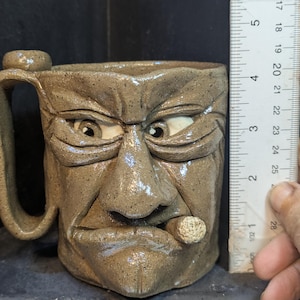 May include: A handmade ceramic mug with a sculpted face design. The mug is approximately 4 inches tall, featuring a handle and a detailed face with a cigar in the mouth. The mug is a neutral tan color.