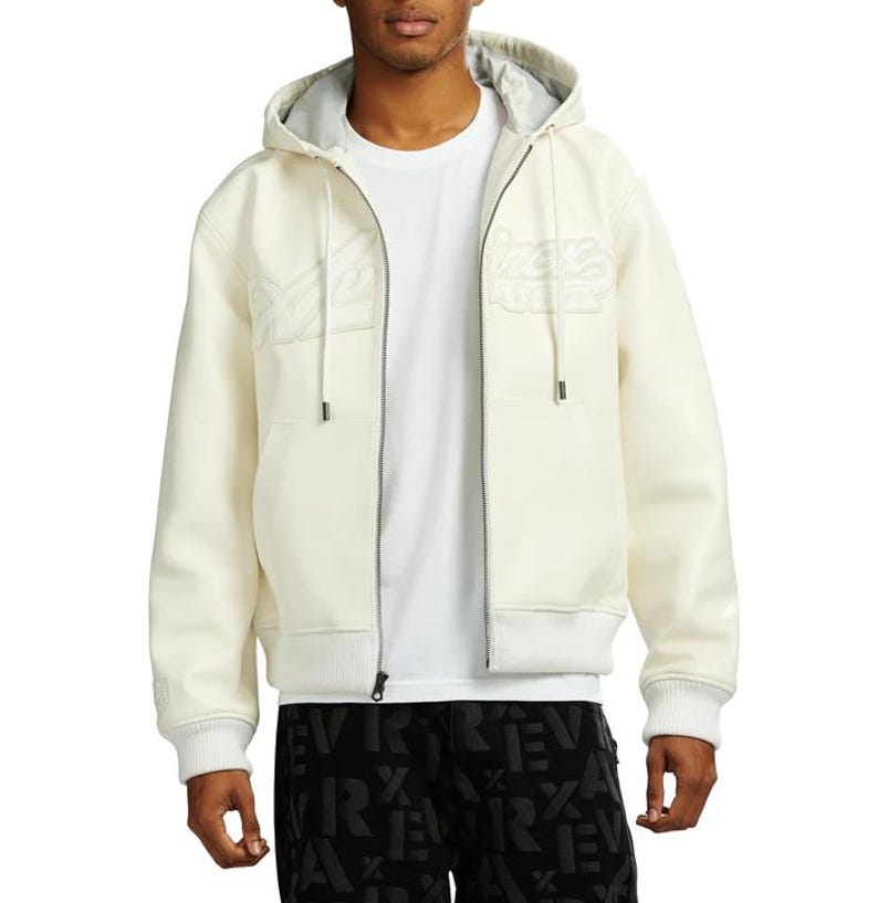 May include: Cream-colored zip-up hoodie with a gray interior lining. The jacket features embroidered text on the chest and a ribbed hem and cuffs. The model is wearing a white t-shirt underneath.