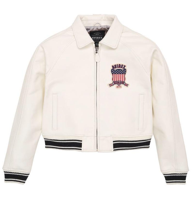 Puede incluir: Chaqueta de cuero color crema con cuello y cremallera. Presenta un parche con un dise&ntilde;o de bandera americana y la marca "AVIREX". La chaqueta tiene pu&ntilde;os y dobladillo a rayas blancas y negras.