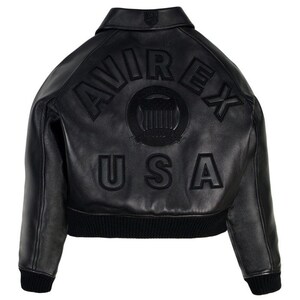 Puede incluir: Chaqueta de cuero negra con la palabra "AVIREX" arqueada en la espalda, encima de un emblema circular y la palabra "USA". La chaqueta tiene dobladillo y pu&ntilde;os de canal&eacute;, y un cuello cl&aacute;sico.