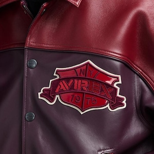 K&ouml;nnte beinhalten: Nahaufnahme einer weinroten und kastanienbraunen Lederjacke. Die Jacke hat einen gro&szlig;en, gestickten Aufn&auml;her mit dem Wort "AVIREX" und dem Jahr "1975". Die Jacke hat einen Knopfverschluss und einen Kragen.