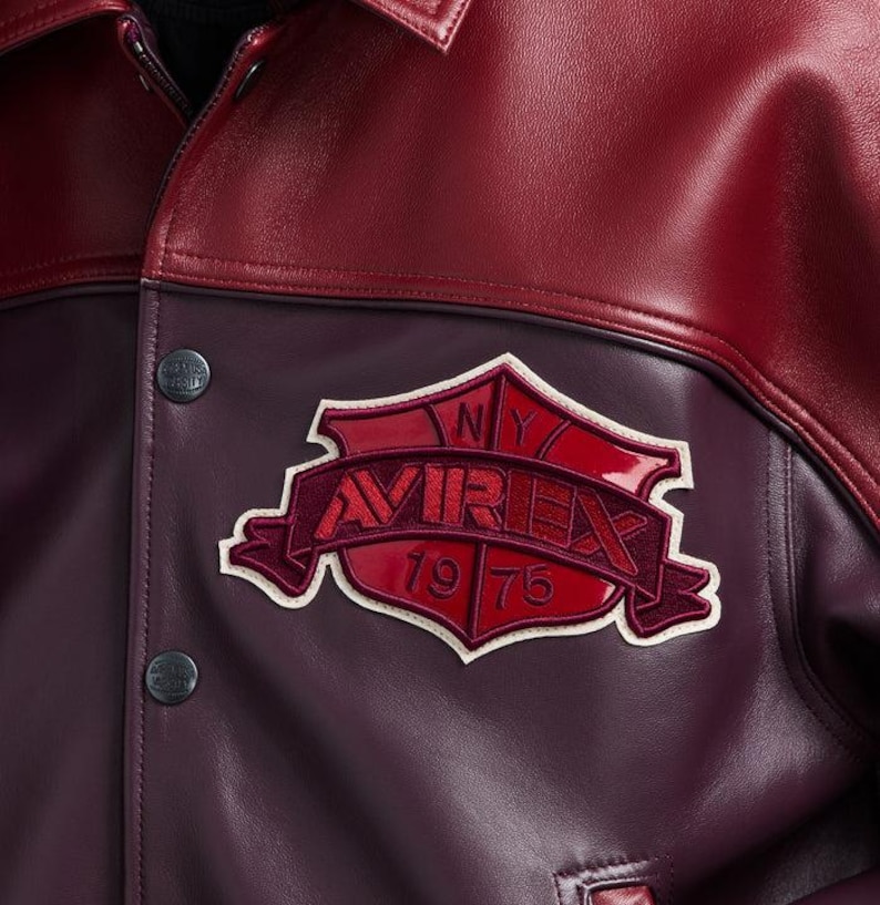 K&ouml;nnte beinhalten: Nahaufnahme einer weinroten und kastanienbraunen Lederjacke. Die Jacke hat einen gro&szlig;en, gestickten Aufn&auml;her mit dem Wort "AVIREX" und dem Jahr "1975". Die Jacke hat einen Knopfverschluss und einen Kragen.