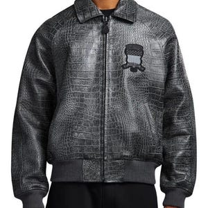 Op de afbeelding: Een grijze leren jas met krokodillenstructuur, een zwarte ritssluiting en een kleine patch op de borst. De jas heeft een bomber-stijl ontwerp met een geribbelde zoom en manchetten. De patch heeft een schildontwerp met een vlag.