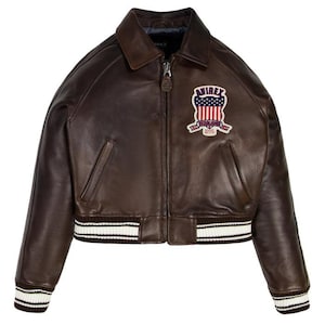 Puede incluir: Chaqueta bomber de cuero marr&oacute;n con cremallera, cuello y parche en el pecho. El parche presenta un dise&ntilde;o rojo, blanco y azul con el texto "AVIREX ESTABLISHED 1975". La chaqueta tiene pu&ntilde;os y dobladillo a rayas blancas y negras.