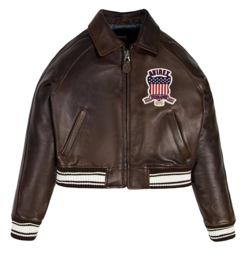 Puede incluir: Chaqueta bomber de cuero marr&oacute;n con cremallera, cuello y parche en el pecho. El parche presenta un dise&ntilde;o rojo, blanco y azul con el texto "AVIREX ESTABLISHED 1975". La chaqueta tiene pu&ntilde;os y dobladillo a rayas blancas y negras.