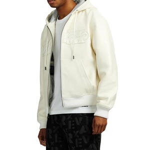May include: Cream-colored zip-up hoodie with a drawstring hood and front pockets. The hoodie features a tonal embroidered design on the chest. The model is wearing a white t-shirt underneath. The bottom of the hoodie has a ribbed hem.