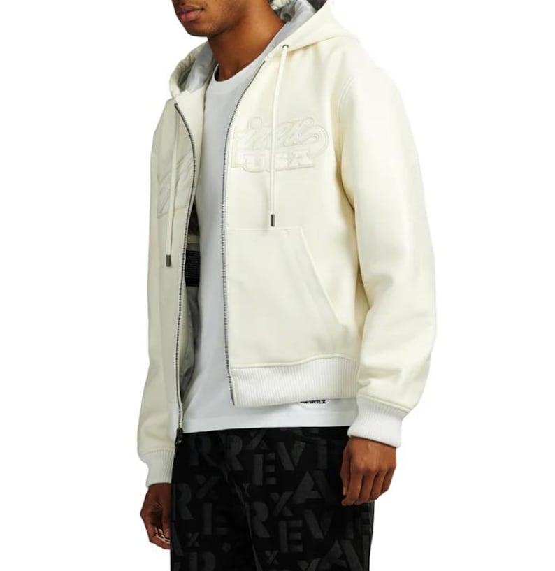 May include: Cream-colored zip-up hoodie with a drawstring hood and front pockets. The hoodie features a tonal embroidered design on the chest. The model is wearing a white t-shirt underneath. The bottom of the hoodie has a ribbed hem.