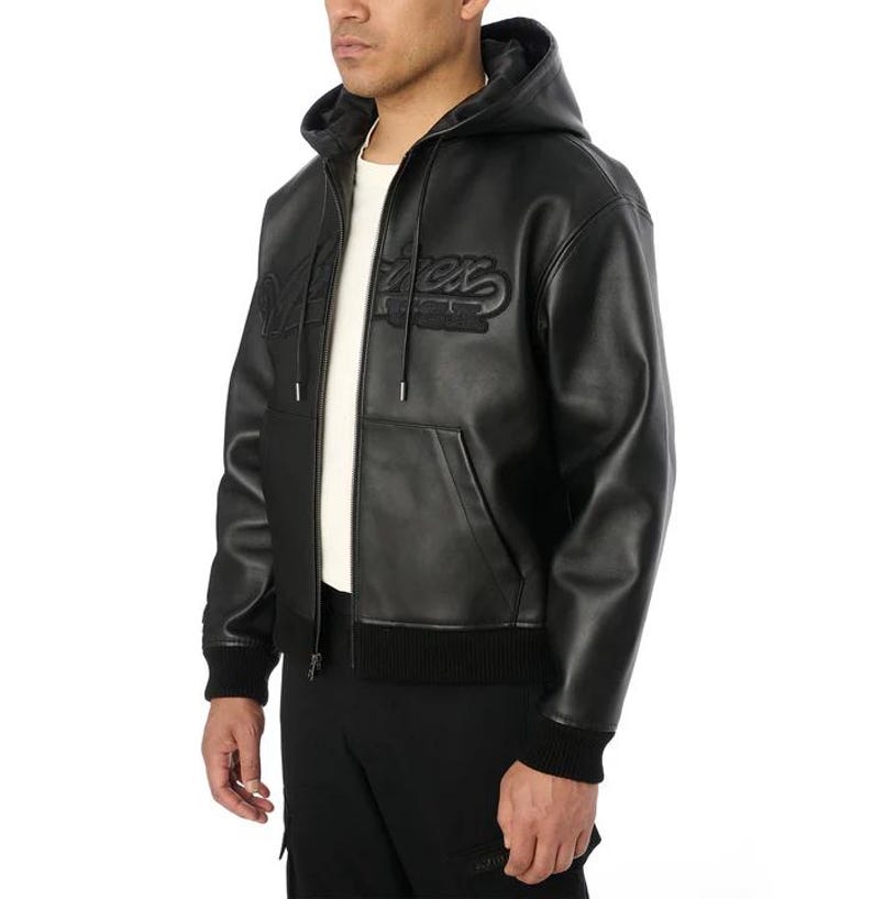 May include: Black leather hooded jacket with a zip-up front, featuring the word "Merx" embroidered on the chest. The jacket has a ribbed hem and cuffs, and a drawstring hood. The model is wearing a white t-shirt underneath.
