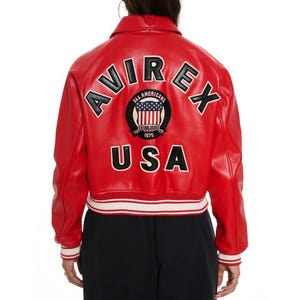 May include: Red leather jacket with white and black accents. The back features the word "AVIREX" in black lettering, along with a patch that says "ALL AMERICANS" and "USA". The jacket has a ribbed white and black hem and cuffs.
