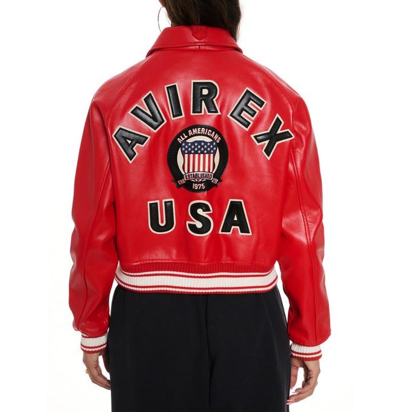 May include: Red leather jacket with white and black accents. The back features the word "AVIREX" in black lettering, along with a patch that says "ALL AMERICANS" and "USA". The jacket has a ribbed white and black hem and cuffs.
