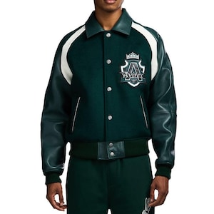 May include: A dark green varsity jacket with white accents and leather sleeves. The jacket features a button-up front, a ribbed hem, and a collar. An emblem with the text "Avirex" is on the chest. The jacket is paired with dark green pants.