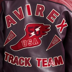 K&ouml;nnte beinhalten: Burgunderfarbene Lederjacke mit roten Akzenten. Auf der R&uuml;ckseite steht das Wort "AVIREX" in wei&szlig;er und roter Schrift, ein "50 USA"-Patch und der Text "TRACK TEAM". Patches zeigen die Jahreszahlen 1975 und 2025.