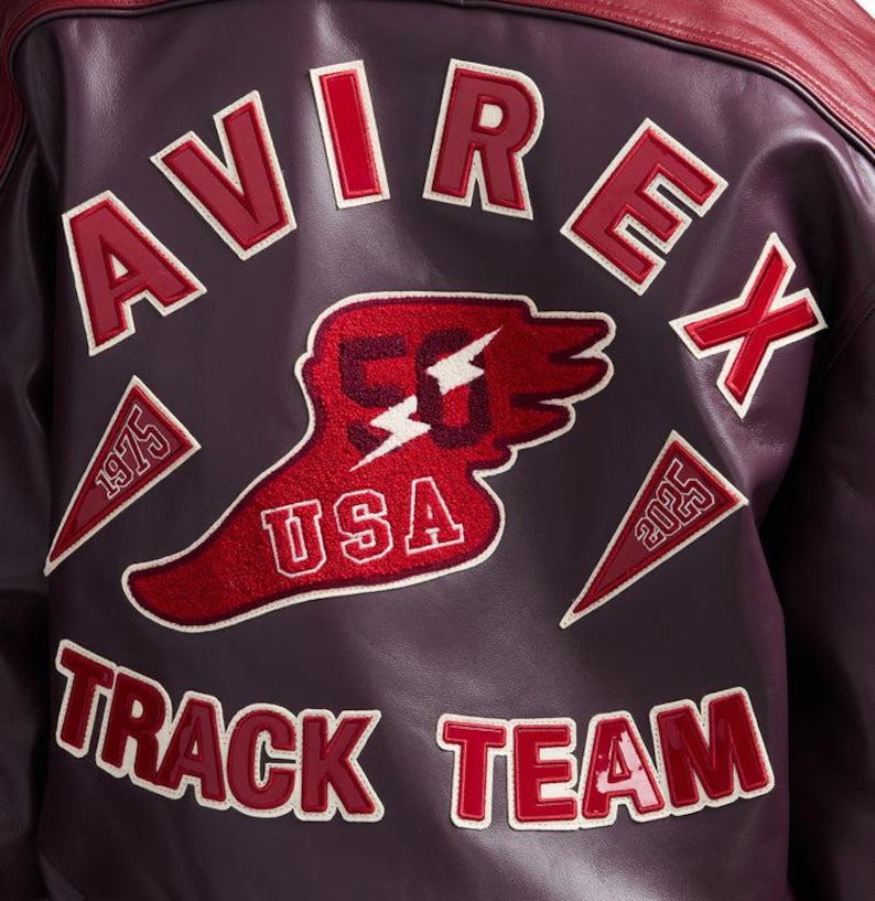 K&ouml;nnte beinhalten: Burgunderfarbene Lederjacke mit roten Akzenten. Auf der R&uuml;ckseite steht das Wort "AVIREX" in wei&szlig;er und roter Schrift, ein "50 USA"-Patch und der Text "TRACK TEAM". Patches zeigen die Jahreszahlen 1975 und 2025.