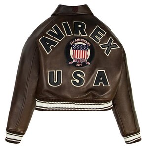 Puede incluir: Chaqueta de cuero marr&oacute;n con las palabras "AVIREX" y "USA" en letras blancas. Un parche con la bandera estadounidense y las palabras "ALL AMERICANS ESTABLISHED 1975" est&aacute; centrado en la espalda. La chaqueta tiene un dobladillo y pu&ntilde;os a rayas blancas y marrones.