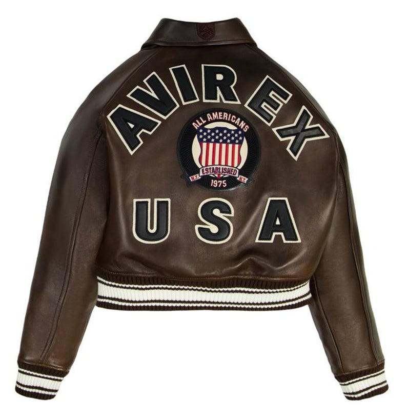 Puede incluir: Chaqueta de cuero marr&oacute;n con las palabras "AVIREX" y "USA" en letras blancas. Un parche con la bandera estadounidense y las palabras "ALL AMERICANS ESTABLISHED 1975" est&aacute; centrado en la espalda. La chaqueta tiene un dobladillo y pu&ntilde;os a rayas blancas y marrones.