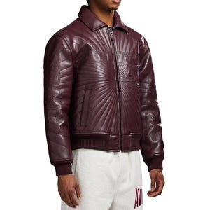 May include: A burgundy leather jacket with a zip-up front, collar, and ribbed cuffs and hem. The jacket features a sunburst design and the word "AVIREX" across the chest. A side pocket is visible.