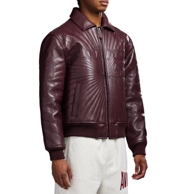 May include: A burgundy leather jacket with a zip-up front, collar, and ribbed cuffs and hem. The jacket features a sunburst design and the word "AVIREX" across the chest. A side pocket is visible.