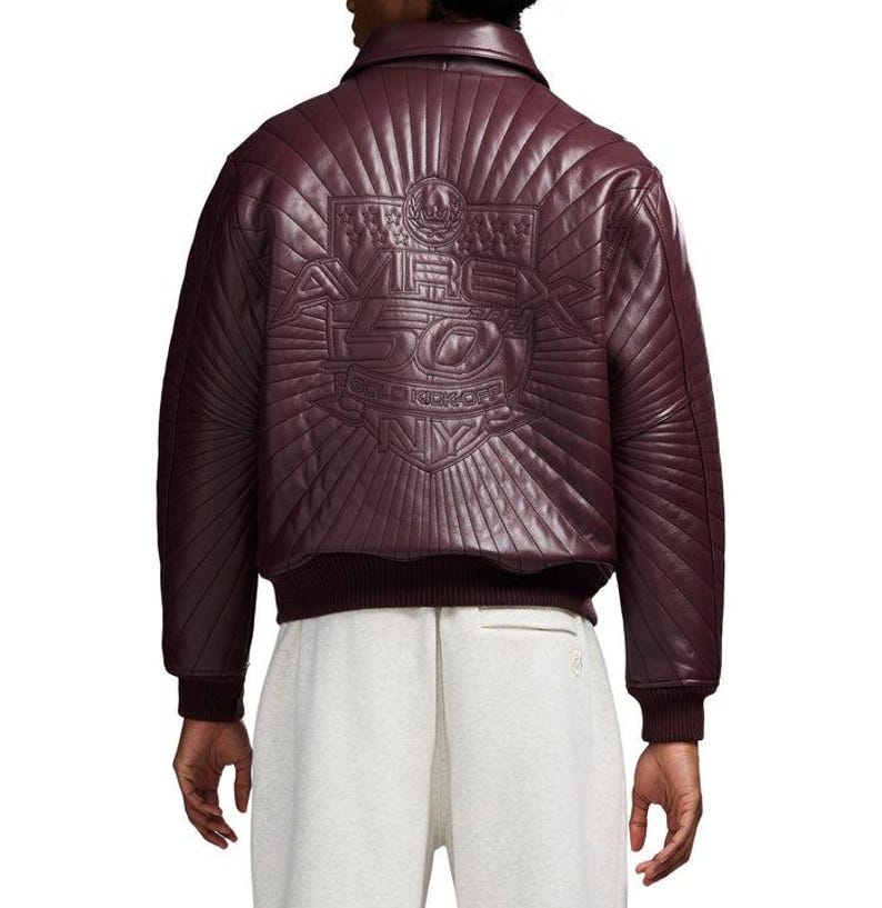 May include: Burgundy leather bomber jacket with quilted back panel. The back features an embossed design with the text "AVE 50 GOLD KICKOFF NY". The jacket has a ribbed hem and cuffs.