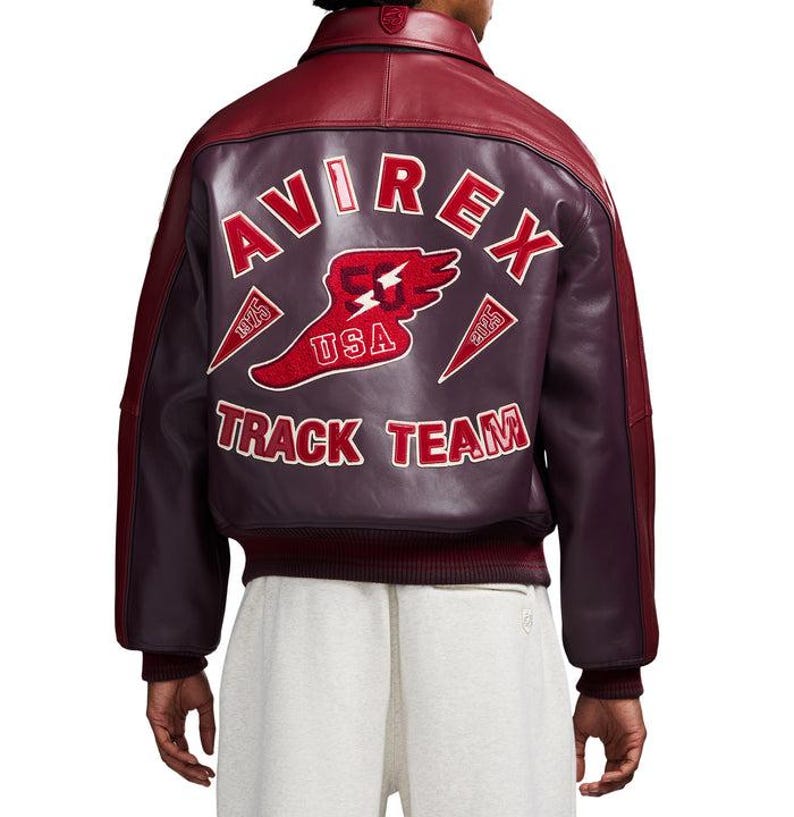 K&ouml;nnte beinhalten: Burgunderrote und dunkelviolette Lederjacke mit dem Text "AVIREX TRACK TEAM" auf dem R&uuml;cken. Die Jacke hat ein rot-wei&szlig;es Logo mit einem gefl&uuml;gelten Schuh und dem Text "USA". Sie hat einen gerippten Saum und B&uuml;ndchen.