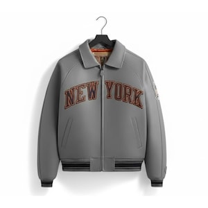 Op de afbeelding: Een grijze bomberjack met de tekst "NEW YORK" in bruine en oranje letters over de borst. De jas heeft een ritssluiting aan de voorkant, zijzakken en zwart-witte strepen op de manchetten en zoom. De jas hangt aan een zwarte hanger.