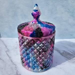 Puede incluir: Un tarro decorativo con tapa, con un patrón de diamantes en el cuerpo. La tapa y la parte superior del tarro están arremolinadas con colores rosa, azul, negro y plateado. El tarro tiene un remate puntiagudo en la parte superior.