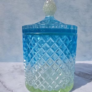 Puede incluir: Un tarro decorativo con tapa, con un patrón de diamantes. El tarro hace una transición de azul a un tono verde claro en la base. La tapa tiene un remate transparente y texturizado. El tarro es probablemente de vidrio o material similar.