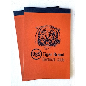 May include: Two orange notebooks with a black tiger illustration and the text "(USS) Tiger Brand Electrical Cable." The notebooks have a dark blue border at the top.