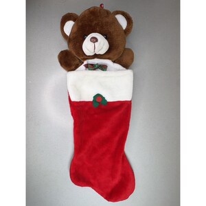 May include: A Christmas stocking with a brown teddy bear peeking out. The stocking is red with a white cuff and a green and red felt decoration. The bear has a white face and ears, and a small bow tie.