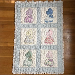 May include: A rectangular quilted blanket featuring six square panels, each displaying a sunbonnet-style figure in different pastel colors including lavender, blue, yellow, pink, green, and mauve. The quilt has a light blue border and ruffled edges.