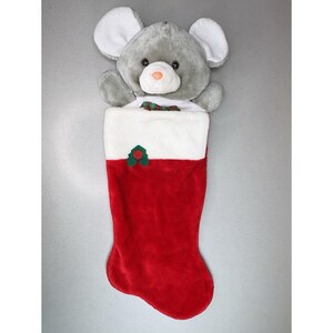 May include: A plush Christmas stocking with a gray mouse head peeking out. The stocking is red with a white cuff and a green holly accent. The mouse has large white ears and a pink nose.