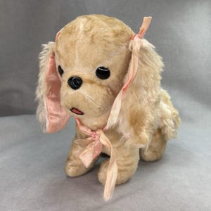 May include: A vintage plush toy dog in a seated position. The dog has tan fur, black eyes, and a black nose. Pink ribbons adorn its ears and neck, adding a touch of color. The toy is made of soft, plush material.
