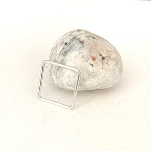 May include: A silver square ring with a smooth, polished finish. The ring is displayed on a white, speckled rock.