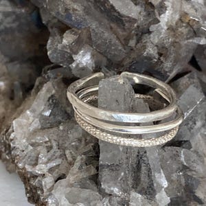 May include: A stackable silver ring set. The rings have different textures, including a hammered finish and a beaded band. The rings are displayed on a rough, gray crystal surface.