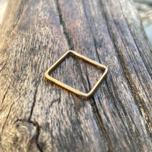 Könnte beinhalten: Ein goldfarbener quadratischer Ring. Der Ring hat ein einfaches, geometrisches Design mit quadratischer Form. Der Ring liegt auf einem rustikalen Holzgrund.