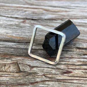May include: A square-shaped silver ring with a smooth, polished finish. The ring is positioned around a dark, elongated, faceted black stone that rests within the center of the square frame, showcasing a contrast between the metal and stone.