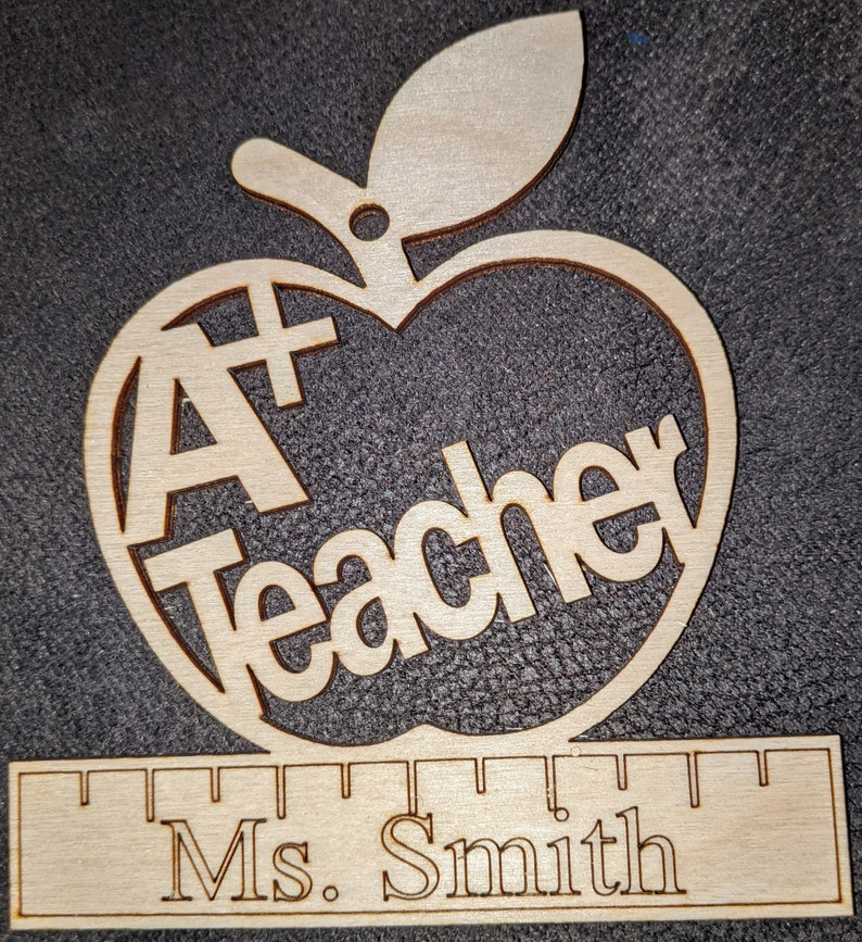 Pu&ograve; includere: Ciondolo a forma di mela in legno con le parole "A+ Teacher" e un righello con il nome "Ms. Smith" inciso su di esso.