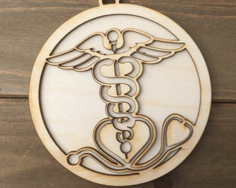 Può includere: Un ornamento in legno a forma di cerchio con un design ritagliato del simbolo medico, il bastone di Asclepio, con uno stetoscopio avvolto attorno alla parte inferiore.