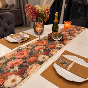 May include: A dining table setting featuring a floral table runner with cream lace trim, mustard yellow placemats, white scalloped plates, clear glass coupes, orange tulip-shaped candle holders, and a bottle of sparkling wine with a bouquet.
