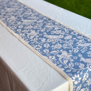 Può includere: Runner da tavolo blu con un motivo floreale e animale bianco, con uccelli e conigli. Ha un bordo in pizzo color crema ed è appoggiato su una tovaglia bianca. Tessuto per la casa decorativo.