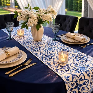 Könnte beinhalten: Ein elegant gedeckter Esstisch mit einer marineblauen Tischdecke und einem Tischläufer mit blau-weißem Blumenmuster. Der Tisch ist mit Tellern, goldenem Besteck und blauen Gläsern gedeckt. Eine Vase mit weißen Blumen und Kerzen vervollständigen die Szene.