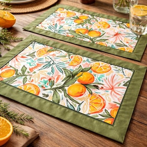 May include: Two rectangular placemats with a white background, featuring a watercolor design of oranges, leaves, and flowers. The placemats have a green border. The scene is set on a wooden table with other dining accessories.