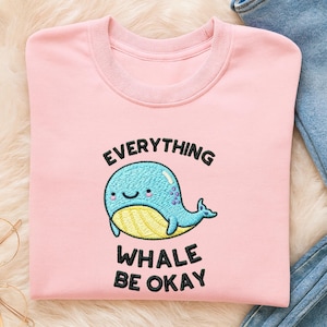 May include: A light pink sweatshirt with the embroidered phrase "EVERYTHING WHALE BE OKAY" and a cartoon whale. The whale is blue with a yellow belly and a smiling face. The sweatshirt has a crew neck and long sleeves.