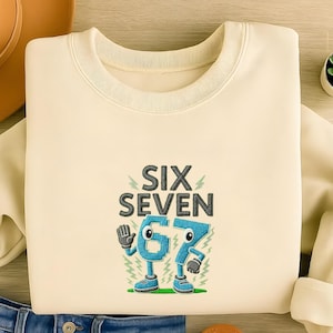 以下が含まれることがあります： クリーム色のスウェットシャツ。グレーで「SIX SEVEN」と刺繍され、青色の漫画風「67」が描かれています。数字には腕、足、靴があり、下には緑の草があります。デザインは前面の中央に配置されています。