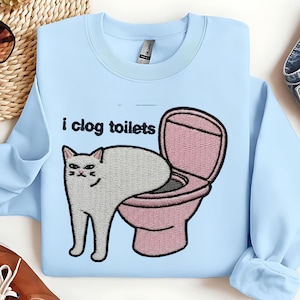 May include: Light blue sweatshirt with an embroidered design featuring a white cat and a pink toilet. The text "i clog toilets" is above the cat and toilet illustration. The sweatshirt has a crew neck and long sleeves.