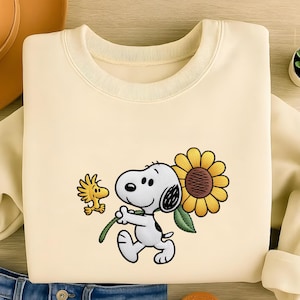 May include: Cream-coloured sweatshirt featuring a cartoon Snoopy character holding a sunflower with Woodstock. The sunflower has yellow petals and a brown centre. The design is embroidered on the front of the sweatshirt.