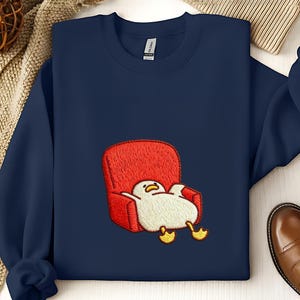 May include: A navy blue sweatshirt with an embroidered design. The design features a cartoon duck relaxing in a red armchair. The duck is white with yellow feet and a yellow beak. A comfortable, casual garment.
